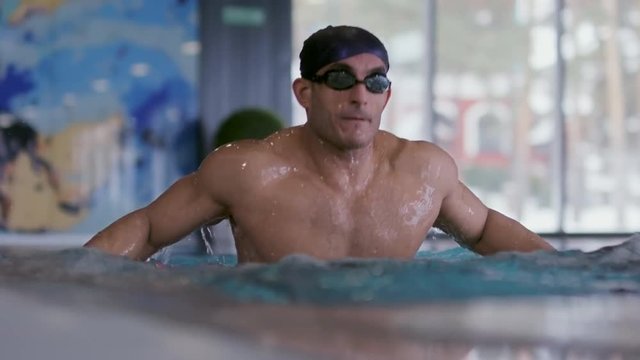 Medium shot of muscular swimmer in cap and goggles jumping out of water in indoor pool, and then walking away in slow motion