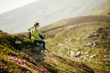 Woman sitting on cliff enjoying mountains landscape. Shot of a young woman looking at the landscape while hiking in the mountains. Magic pink rhododendron flowers on summer mountain
