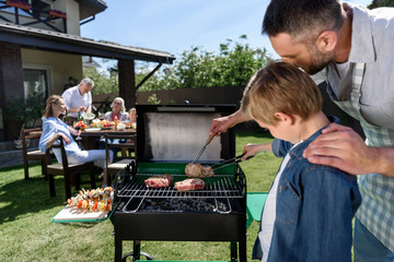 Close-up view of father and son grilling meat while family sitting at table outdoors