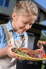 smiling kid girl holding plate with fresh cooked steak outdoors