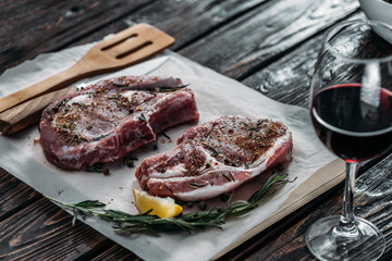 Glass of red wine with raw ribeye steaks decorated by rosemary, spices and lemon slice on baking paper