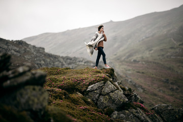Naklejka na ściany i meble Young hipster woman hiker hiking on mountain trail. Travel concept. Traveler standing on top of a mountain. Hiker girl wrapping in warm poncho outdoor. Beautiful boho woman
