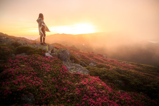 young woman hiking at mountain peak above clouds and fog. Hiker girl wrapping in warm poncho outdoor. Young girl over the clouds. Wonderful landscape with cloud inversion. Flowers in the mountains
