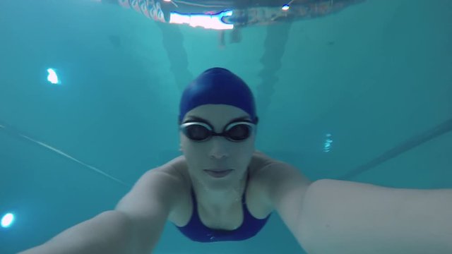 Female athlete in cap and goggles holding camera and swimming underwater in indoors pool, then emerging 