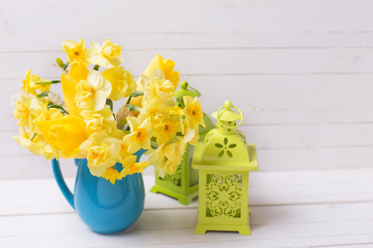 Spring daffodils or narcissus flowers in  blue pitcher and decorative green lanterns on white  wooden background.