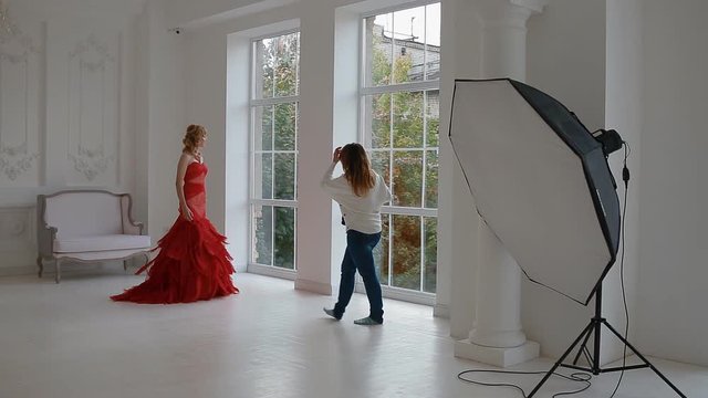 Female photographer work with model in studio. Backstage. Young woman in luxury evening dress posing in white interior.