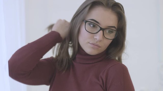 Portrait of attractive young woman in the red sweater with glasses standing near window, turning, possing and smiling on camera.