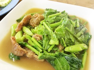 Stir-fried kale vegetable with crispy pork in white dish on the table. Thai street food.