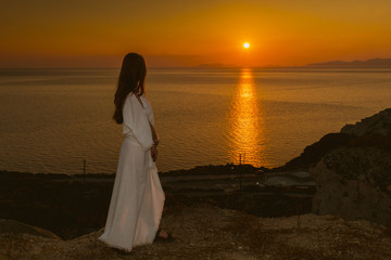 Young girl on the edge of a cliff in a white dress watching the sunset in Santorini, Greece. Female in white sundress on summer Europe travel vacation.