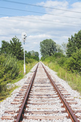 Fototapeta premium Rusted railroad tracks and sleepers on white gravel, surrounded with greenery and blue sky with white clouds on a summer day