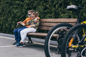 stylish kids in sunglasses embracing while resting on bench at park