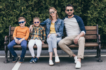 smiling family in sunglasses sitting together on bench at park