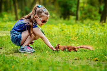 A little girl in denim overalls, a T-shirt, sneakers and glasses feeds a small red squirrel from her hand. - 162233872
