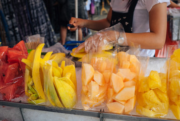 Street fruit vendor is selling assorted kinds of fruits, cantaloupe,  red water melon, yellow mango, pineapple. The vendor is slicing fruit with a knife and put pieces into plastic bags. 