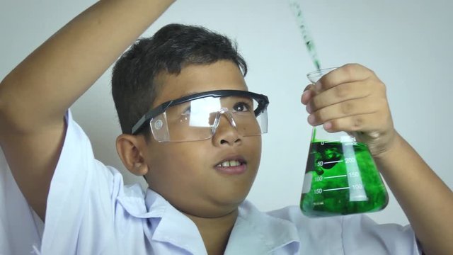 Medium close ethnic boy studying a green liquid chemical solution in a beaker as part of S.T.E.M  science education. He holds the bottle close to his eye and looks inside - then makes a funny face