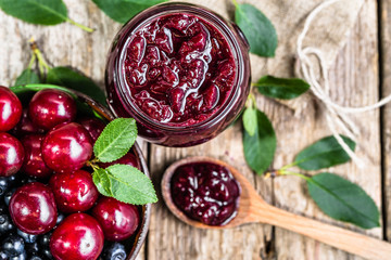 Cherry jam in jar and fresh cherries in a bowl, homemade preserves on rustic background - obrazy, fototapety, plakaty