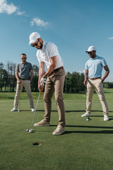 Young golfer putting ball into hole while his friends standing behind