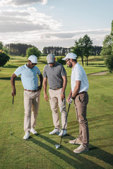 Multiethnic group of golfers holding clubs and talking while standing on green grass