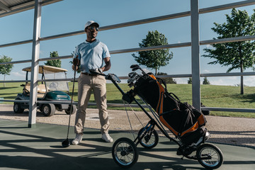 stylish golf player with golf club in hand standing near golf bag with equipment