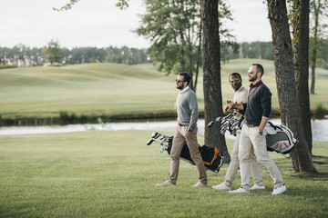 Multiethnic golf players holding bags with golf clubs and walking on golf course