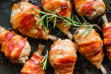 Oven roasted bacon wrapped chicken drumsticks in a black baking pan, top view.