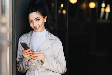 Fototapeta na wymiar Portrait of adorable bruntte woman with dark eyes and eyebrows, pure healthy skin dressed in white clothes holding mobile phone in hands demonstrating her perfect manicure looking into camera