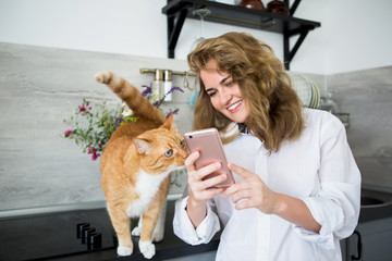 Woman in a white man's shirt is standing in the kitchen. A girl shows the cat a smartphone screen and smiles. Cat is looking at the phone.