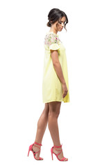 Sophisticated feminine young woman in short yellow evening dress walking and looking down. Full body length portrait isolated on white background. 