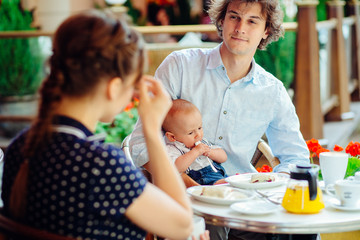 young happy family of mother, father and newborn baby boy having breakfast in the street cafe in summer time
