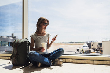 Fototapeta na wymiar Young woman in the airport, using smart phone and drinking coffee, travel, vacations and active lifestyle concept
