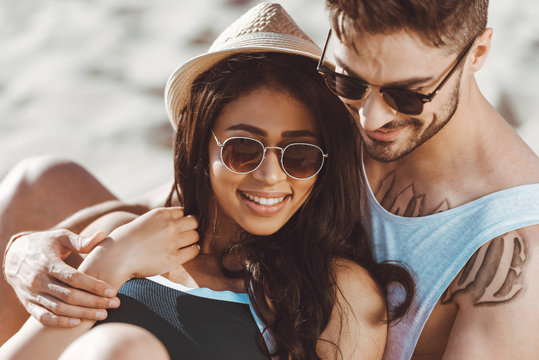Happy young couple in sunglasses sitting and embracing on beach