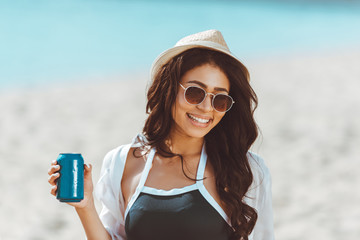 Beautiful young woman in sunglasses and straw hat holding soda can and smiling at camera - Powered by Adobe
