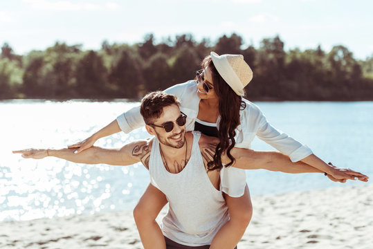 Handsome young man in sunglasses piggybacking smiling girl in straw hat at beach