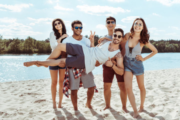 cheerful friends holding man in hands and looking at camera on beach