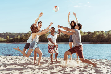 young smiling friends playing beach volleyball on riverside at daytime