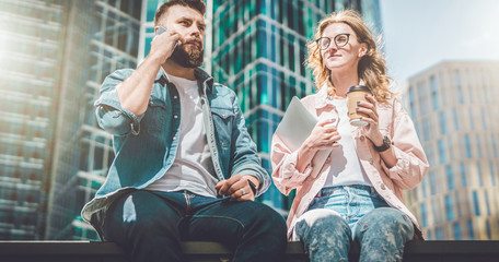 Two young business people are sitting on street. Hipster guy is talking on cell phone, girl is drinking coffee