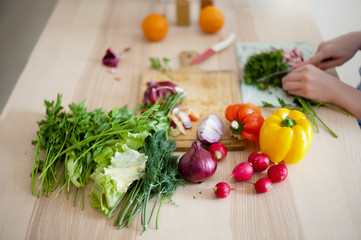 Fresh vegetables and herbs lying on the kitchen table, in the background woman cut vegetables for salad - 162267055