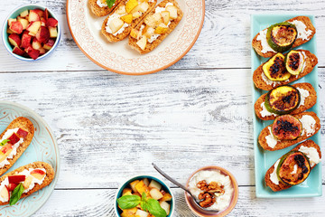 Small toasts (crostini) with cream cheese and cut fresh fruits (pear, peach, cherry, grilled figs) on white table. Top view of summer appetizers with copy space.