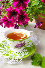 Herbal tea with a slice of lemon and a sprig of aromatic mint on a tablecloth in a rural style and a bouquet of petunia flowers in a vase. The concept of a romantic morning breakfast in the summer