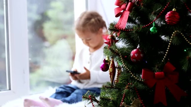 Decorated Christmass tree and little girl with smart phone on the background.