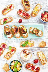 Top view of assorted crostini (toasts) with cut fresh fruits: cherry, peach, pear and grilled figs on white wooden table. Healthy summer picnic snack.