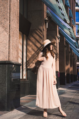 Close-up portrait of a slender young girl blogger beautiful brunette in downtown dusseldorf in a pastel dress and a lady's hat wearing sunglasses walking poses and smiling on the sunset