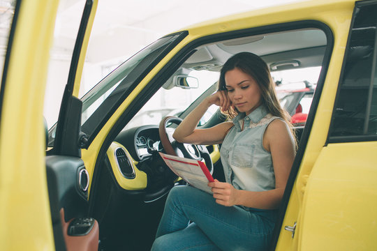 The young dark-haired woman examining car at the dealership and making his choice. Horizontal portrait of a young female model at the car. He is thinking if he should buy it