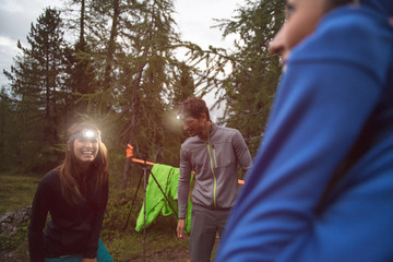 Happy smiling woman and man with headlamp flashlight during evening near camping. Group of friends people summer adventure journey in mountain nature outdoors. Travel exploring Alps, Dolomites, Italy. - obrazy, fototapety, plakaty