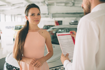 friendly car salesman talking to a young woman and showing a new car inside showroom Signing of contract.