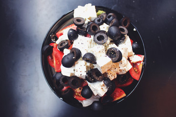 Greek salad with sirtaki cheese and black olives and a red tomato. On white cheese is ground pepper. Salad in a glass plate is on a dark table.