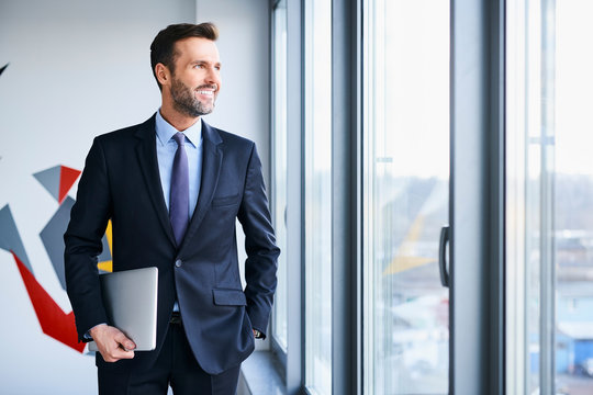 Portrait of middle-aged businessman looking through window while holding laptop
