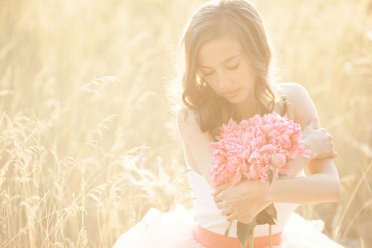 Beautiful young girl with pions on the nature otdoors in the sunshine. Pretty woman outside with flowers in the pink skirt