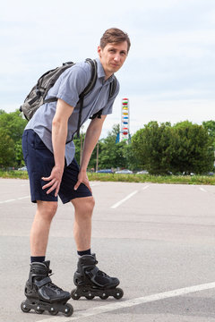 Caucasian man in roller-skates with backpack standing on asphalt road, full-lenght portrait