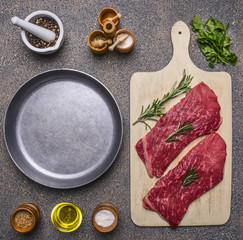two delicious raw beef steak on a cutting board with rosemary and parsley, spices and laid next to the pan, place for text